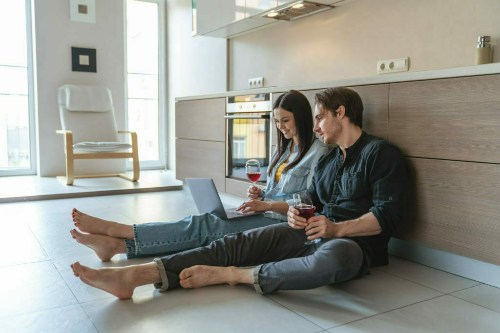 a man and a woman sitting on the floor with a laptop and a glass of wine