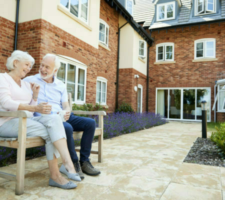 Retired Couple Sitting On Bench With Hot Drink In Assisted Living Facility