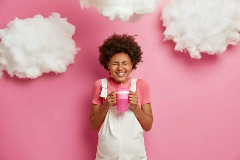 Overjoyed pregnant woman holds bottle with water smiles broadly going to give birth to child dressed in casual clothes has big abdomen poses against pink background. Pregnancy maternity concept